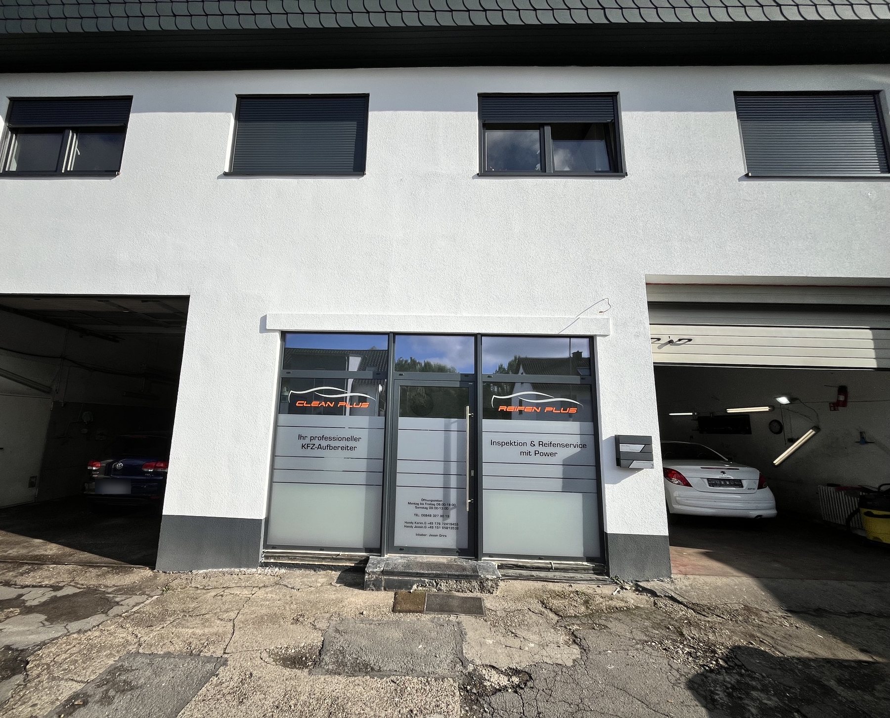 Gebäude von Clean Plus von vorne: weiße Fassade, zwei offene Hallentore und Eingang mit Schriftzug Clean Plus und Reifen Plus